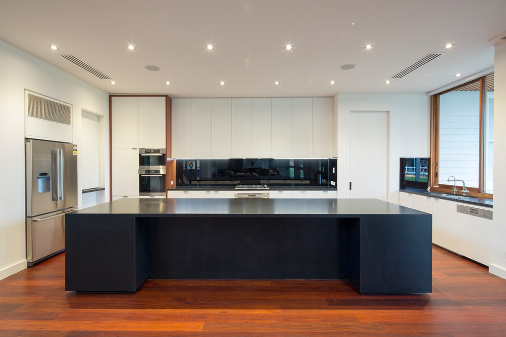 Expansive contemporary open plan kitchen in Perth with an undermount sink, flat-panel cabinets, black cabinets, granite benchtops, black splashback, glass sheet splashback, stainless steel appliances, medium hardwood floors and with island.