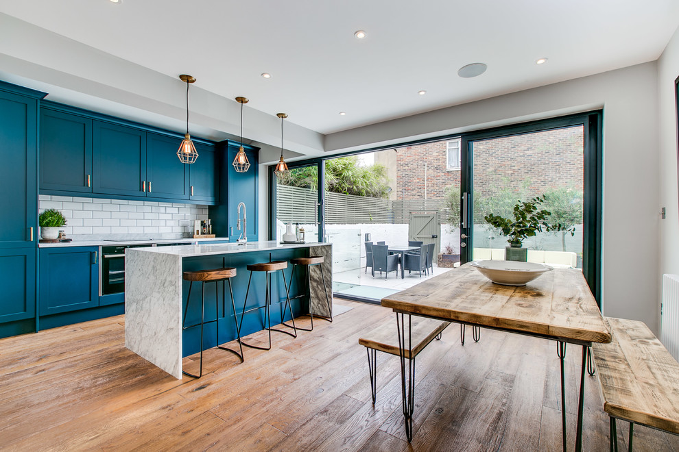Mid-sized contemporary single-wall eat-in kitchen in London with recessed-panel cabinets, blue cabinets, marble benchtops, white splashback, subway tile splashback, stainless steel appliances, light hardwood floors, with island, brown floor and white benchtop.