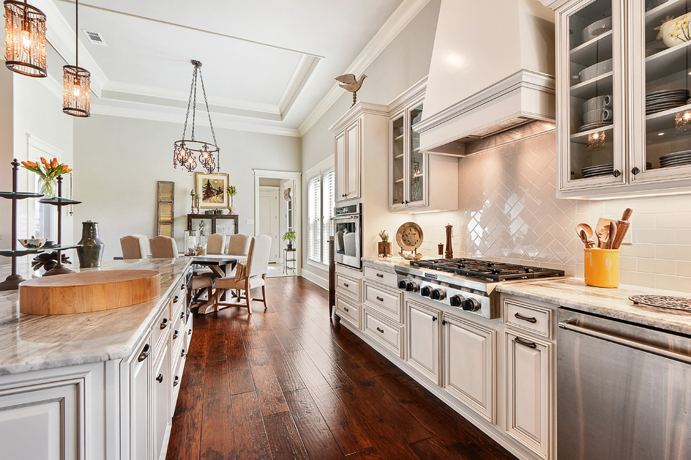 Large transitional l-shaped open plan kitchen in New Orleans with a farmhouse sink, beaded inset cabinets, white cabinets, marble benchtops, beige splashback, ceramic splashback, stainless steel appliances, medium hardwood floors, with island, brown floor and grey benchtop.