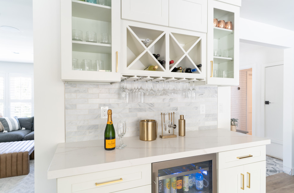 Design ideas for a mid-sized transitional galley home bar in Orange County with no sink, shaker cabinets, white cabinets, quartz benchtops, grey splashback, stone tile splashback, vinyl floors, brown floor and white benchtop.