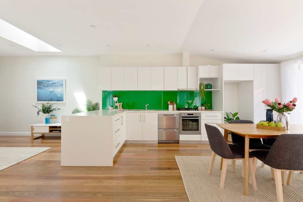 Photo of a contemporary l-shaped eat-in kitchen in Sydney with flat-panel cabinets, white cabinets, green splashback, stainless steel appliances, medium hardwood floors, no island and brown floor.