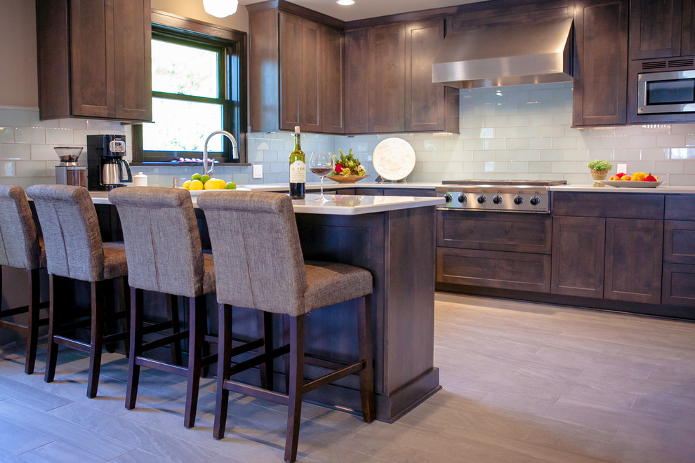 This is an example of a mid-sized transitional l-shaped eat-in kitchen in Portland with shaker cabinets, dark wood cabinets, subway tile splashback, stainless steel appliances, a peninsula, an undermount sink, quartz benchtops, blue splashback and light hardwood floors.