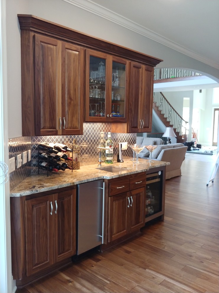 This is an example of a small traditional single-wall wet bar in Milwaukee with an undermount sink, raised-panel cabinets, dark wood cabinets, quartz benchtops, grey splashback, metal splashback, dark hardwood floors, brown floor and beige benchtop.