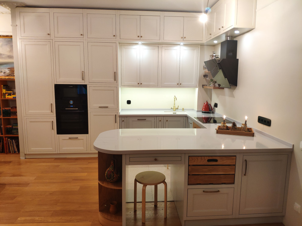 This is an example of a large scandinavian u-shaped eat-in kitchen in Moscow with an undermount sink, raised-panel cabinets, solid surface benchtops, white splashback, black appliances, laminate floors, a peninsula, brown floor and grey benchtop.