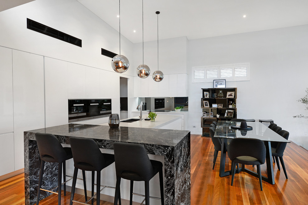 Large contemporary l-shaped eat-in kitchen in Wollongong with a double-bowl sink, flat-panel cabinets, white cabinets, mirror splashback, black appliances, medium hardwood floors, with island, brown floor and white benchtop.