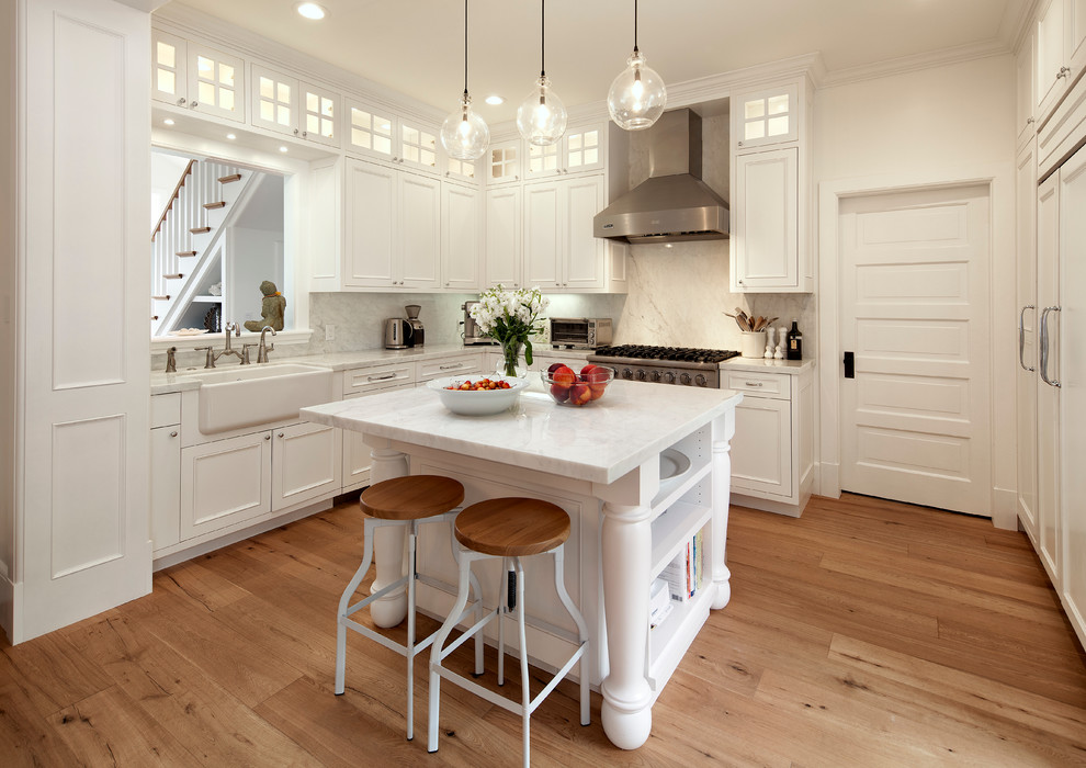 Photo of a mid-sized traditional l-shaped kitchen in Santa Barbara with a farmhouse sink, white cabinets, marble benchtops, white splashback, stone slab splashback, stainless steel appliances, with island, beaded inset cabinets and medium hardwood floors.