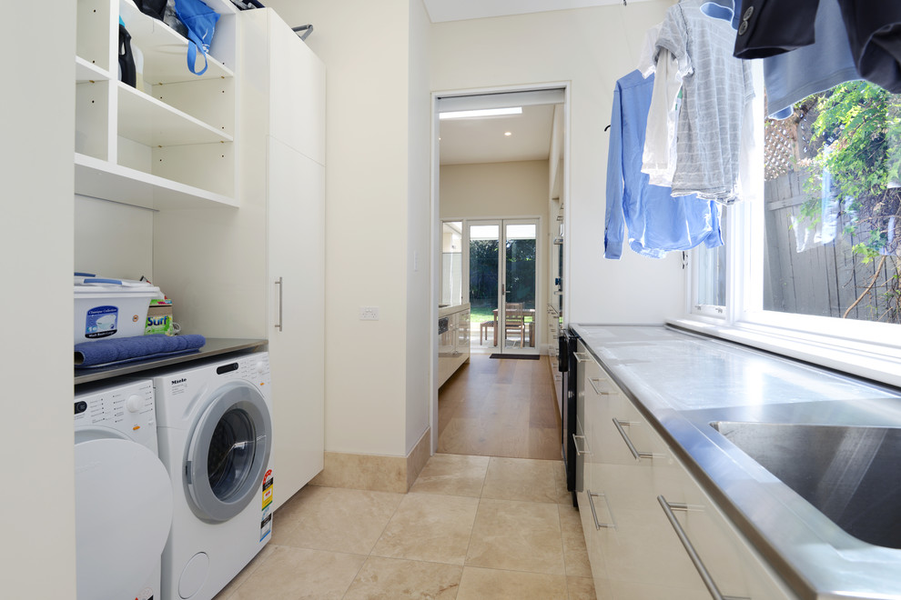 Design ideas for a mid-sized contemporary galley dedicated laundry room in Sydney with a drop-in sink, flat-panel cabinets, white cabinets, stainless steel benchtops, white walls, limestone floors and a side-by-side washer and dryer.