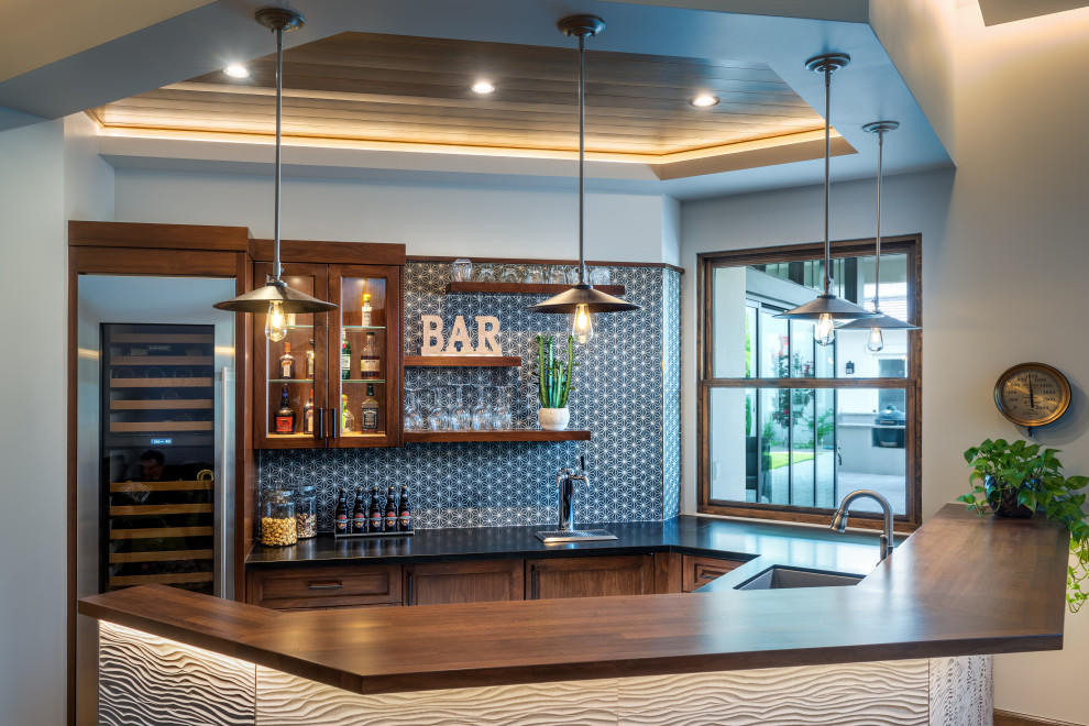 This is an example of a mid-sized country u-shaped wet bar in Phoenix with glass-front cabinets, soapstone benchtops, multi-coloured splashback, mosaic tile splashback, medium hardwood floors, brown floor, black benchtop, an undermount sink and medium wood cabinets.