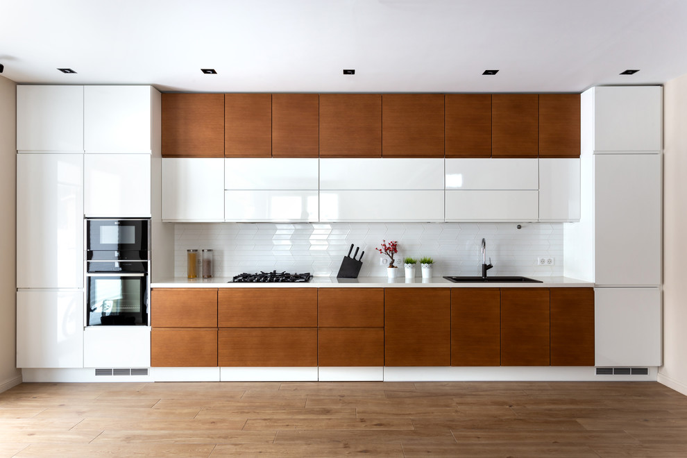 Design ideas for a mid-sized contemporary single-wall eat-in kitchen in Moscow with a drop-in sink, flat-panel cabinets, white cabinets, solid surface benchtops, white splashback, ceramic splashback, black appliances, medium hardwood floors, no island, brown floor and white benchtop.