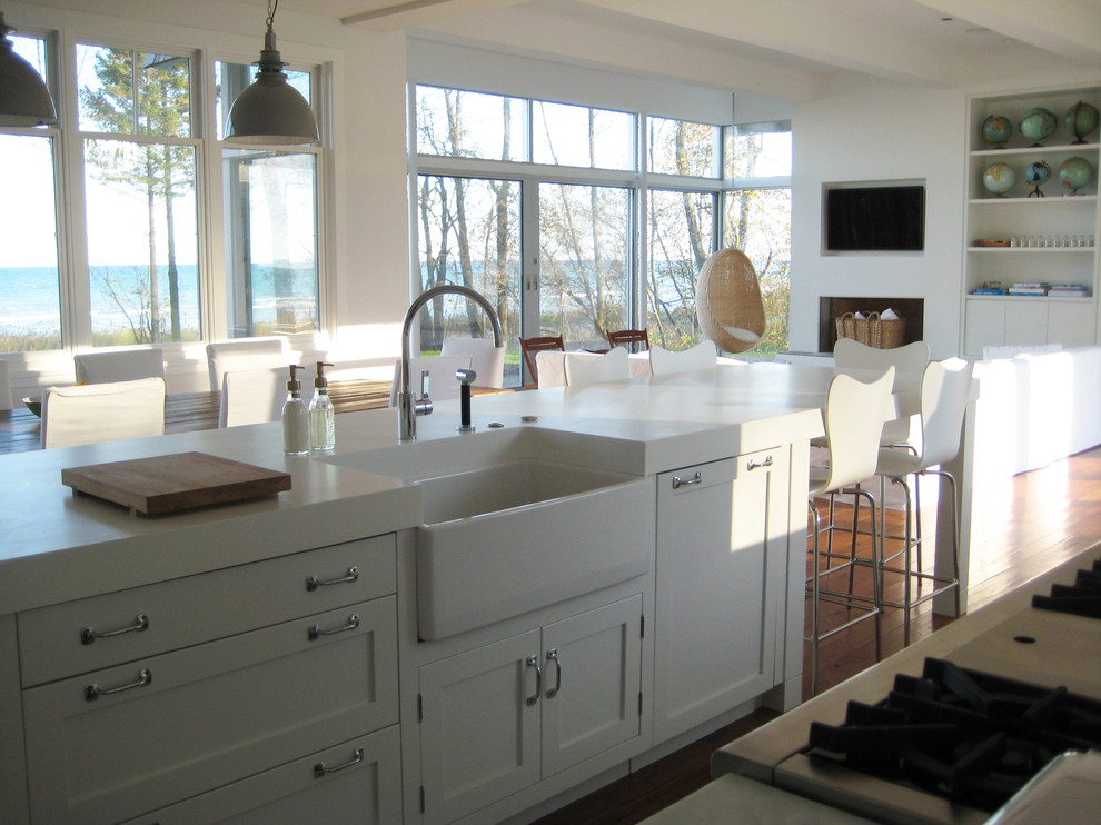This is an example of a beach style galley open plan kitchen in Milwaukee with a farmhouse sink, shaker cabinets, white cabinets, solid surface benchtops, white splashback, stainless steel appliances, medium hardwood floors, with island, brown floor and white benchtop.