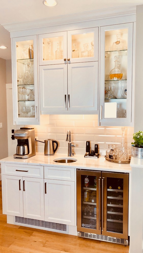 Inspiration for a large contemporary home bar in Other with an undermount sink, shaker cabinets, white cabinets, quartz benchtops, white splashback, subway tile splashback, light hardwood floors, brown floor and white benchtop.