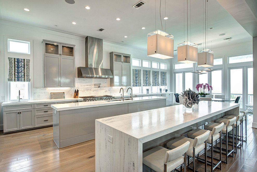 Inspiration for a beach style eat-in kitchen in Houston with a triple-bowl sink, shaker cabinets, grey cabinets, white splashback, stone slab splashback, stainless steel appliances, light hardwood floors, multiple islands and white benchtop.