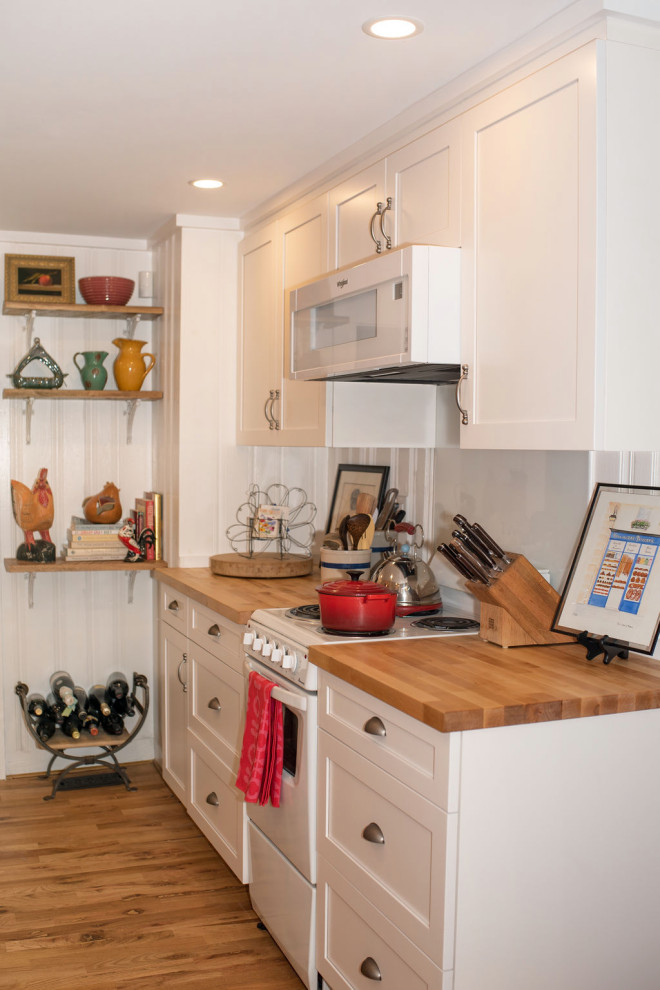 Photo of a small traditional galley separate kitchen in Other with a farmhouse sink, shaker cabinets, white cabinets, wood benchtops, white splashback, glass sheet splashback, white appliances, light hardwood floors, no island, brown floor and brown benchtop.