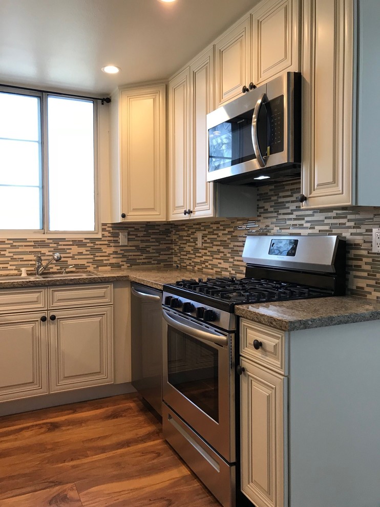 Photo of a small transitional u-shaped separate kitchen in Los Angeles with a drop-in sink, raised-panel cabinets, beige cabinets, granite benchtops, multi-coloured splashback, glass sheet splashback, stainless steel appliances, laminate floors, brown floor and multi-coloured benchtop.