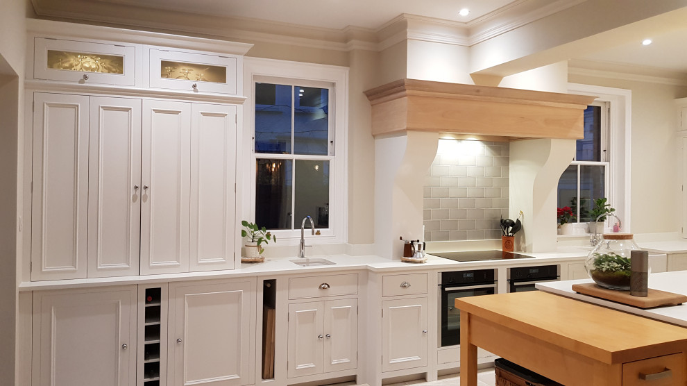 Mid-sized traditional eat-in kitchen in London with a farmhouse sink, beaded inset cabinets, white cabinets, quartzite benchtops, grey splashback, subway tile splashback, black appliances, limestone floors, with island, white floor and white benchtop.