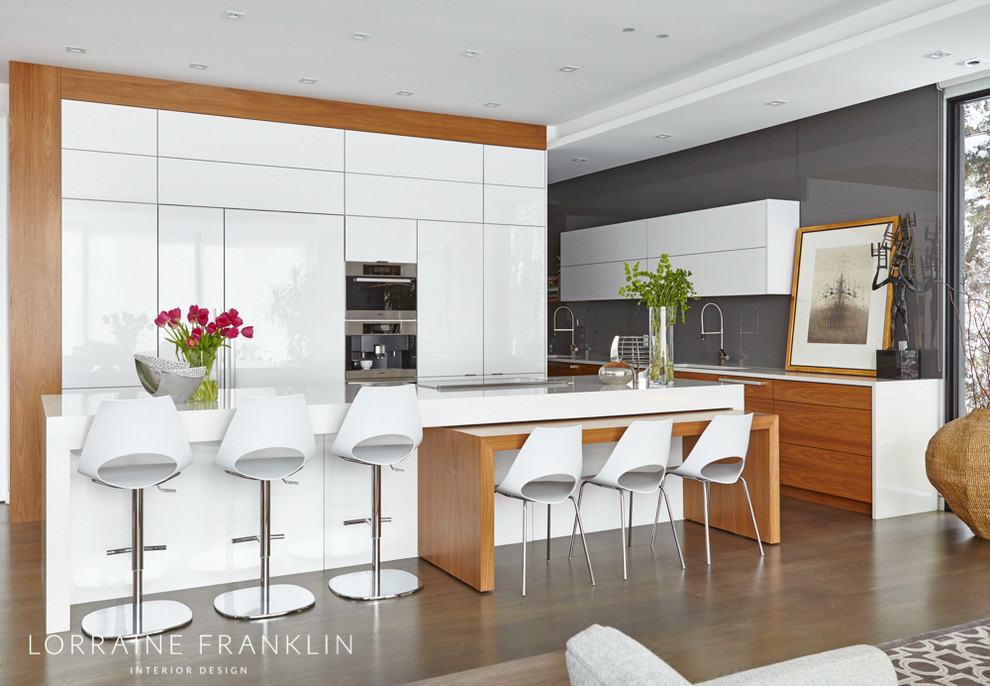 Large modern l-shaped open plan kitchen with white cabinets, grey splashback, glass sheet splashback, panelled appliances, with island, flat-panel cabinets, an undermount sink, quartz benchtops, concrete floors, brown floor and white benchtop.