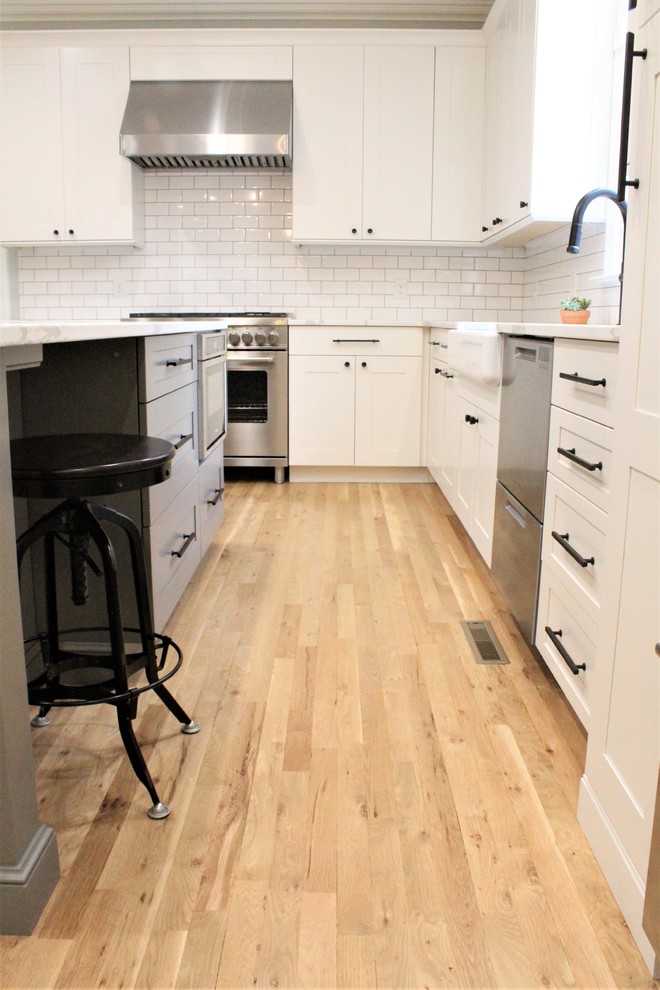 Large transitional galley light wood floor eat-in kitchen photo with a farmhouse sink, shaker cabinets, white cabinets, quartz countertops, white backsplash, ceramic backsplash, stainless steel appliances, an island and white countertops