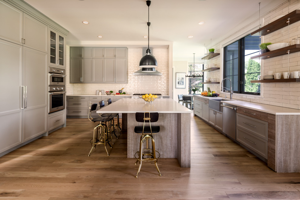This is an example of a country eat-in kitchen in Chicago with a farmhouse sink, shaker cabinets, grey cabinets, quartz benchtops, white splashback, subway tile splashback, stainless steel appliances, light hardwood floors, with island, beige floor and white benchtop.