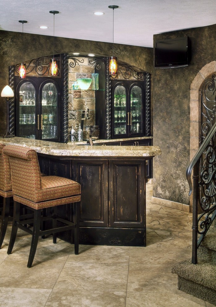 Inspiration for a mid-sized country l-shaped seated home bar in Omaha with raised-panel cabinets, brown cabinets, granite benchtops, travertine floors, beige floor, stone tile splashback, brown benchtop and grey splashback.