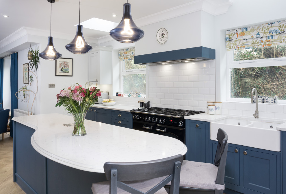 Photo of a large traditional single-wall open plan kitchen in Cardiff with a farmhouse sink, shaker cabinets, blue cabinets, quartz benchtops, white splashback, black appliances, light hardwood floors, with island, brown floor and white benchtop.