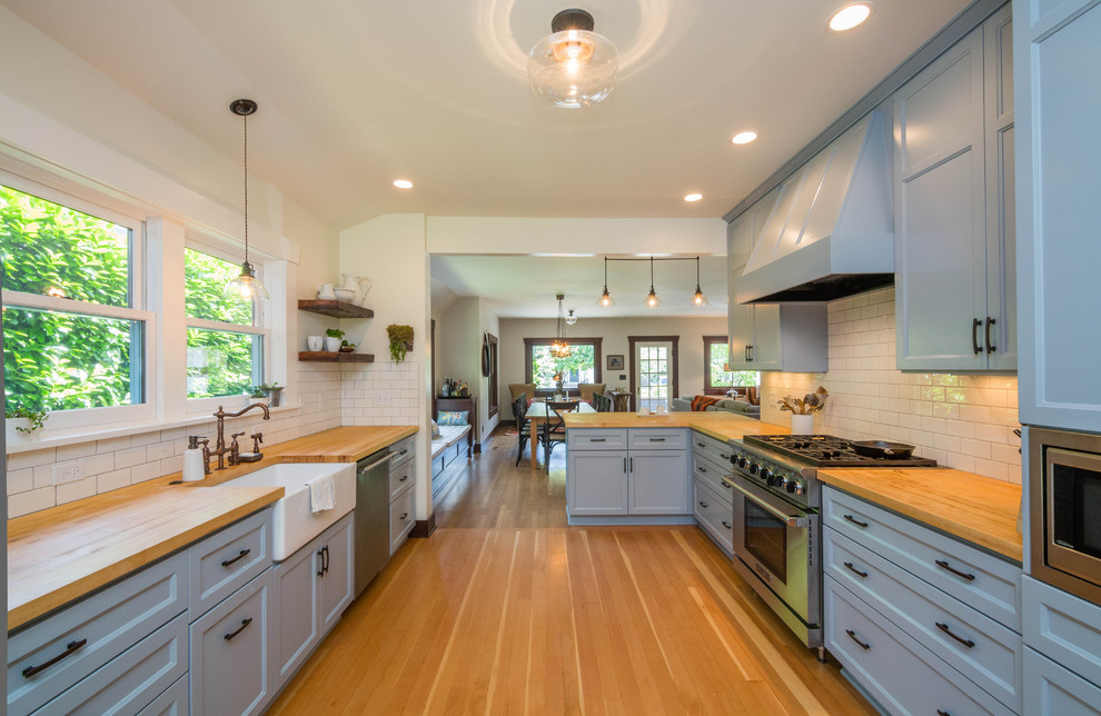 Inspiration for a large transitional u-shaped kitchen in Portland with a farmhouse sink, recessed-panel cabinets, blue cabinets, wood benchtops, white splashback, subway tile splashback, stainless steel appliances, light hardwood floors and a peninsula.
