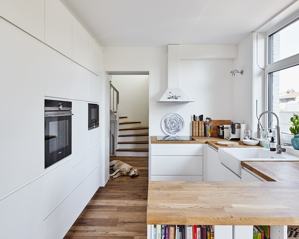 Design ideas for a mid-sized scandinavian u-shaped kitchen in Dusseldorf with a farmhouse sink, flat-panel cabinets, white cabinets, wood benchtops, black appliances, dark hardwood floors and a peninsula.