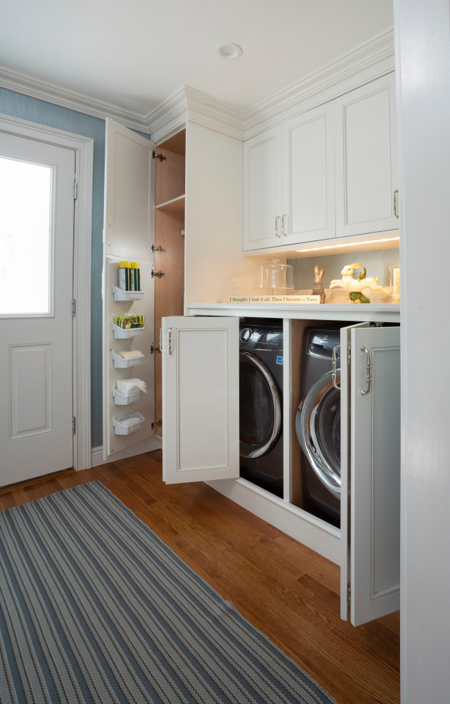 Photo of a mid-sized transitional utility room in New York with white cabinets, blue walls, medium hardwood floors, a side-by-side washer and dryer and brown floor.