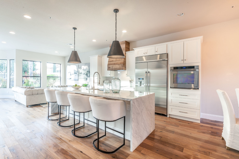 This is an example of a transitional galley open plan kitchen in Phoenix with an undermount sink, shaker cabinets, white cabinets, stainless steel appliances, medium hardwood floors, with island, brown floor and grey benchtop.
