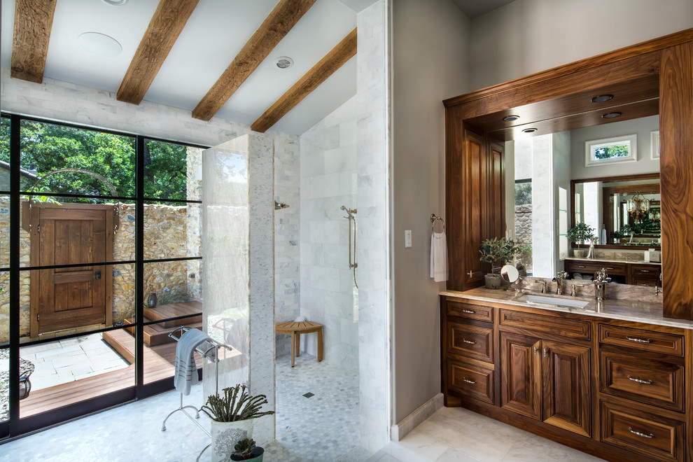 Photo of a mid-sized country master bathroom in Dallas with raised-panel cabinets, dark wood cabinets, a freestanding tub, an alcove shower, a two-piece toilet, beige tile, stone tile, grey walls, marble floors, an undermount sink and marble benchtops.