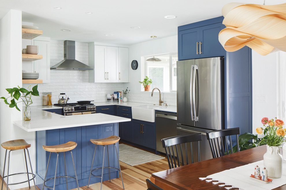 Mid-sized modern u-shaped eat-in kitchen in Austin with a farmhouse sink, shaker cabinets, blue cabinets, quartz benchtops, white splashback, ceramic splashback, stainless steel appliances, light hardwood floors, a peninsula, brown floor and white benchtop.