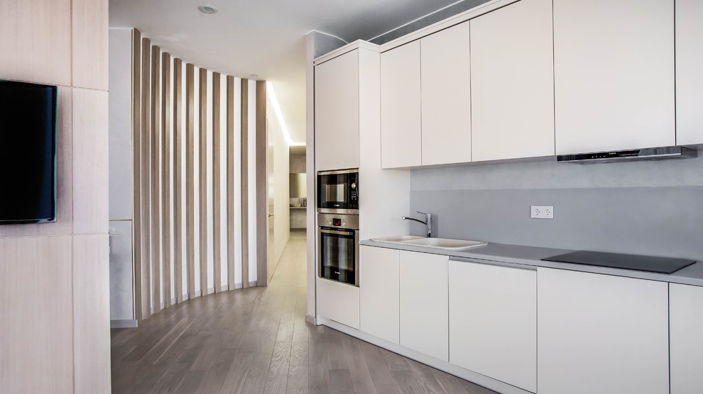 This is an example of a contemporary single-wall open plan kitchen in Moscow with flat-panel cabinets, white cabinets, grey splashback, stainless steel appliances, medium hardwood floors, a double-bowl sink, no island and brown floor.