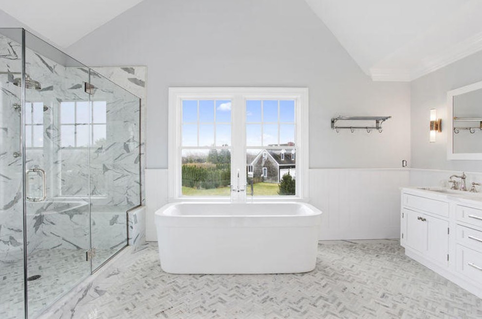 Photo of a large beach style master bathroom in New York with a drop-in sink, beaded inset cabinets, white cabinets, marble benchtops, a freestanding tub, a double shower, white tile, matchstick tile, grey walls and marble floors.