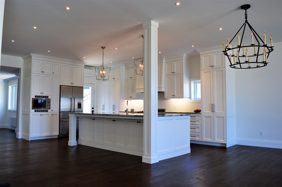 Photo of an expansive traditional l-shaped eat-in kitchen in Toronto with an undermount sink, shaker cabinets, white cabinets, granite benchtops, white splashback, ceramic splashback, stainless steel appliances, dark hardwood floors, with island, brown floor and multi-coloured benchtop.