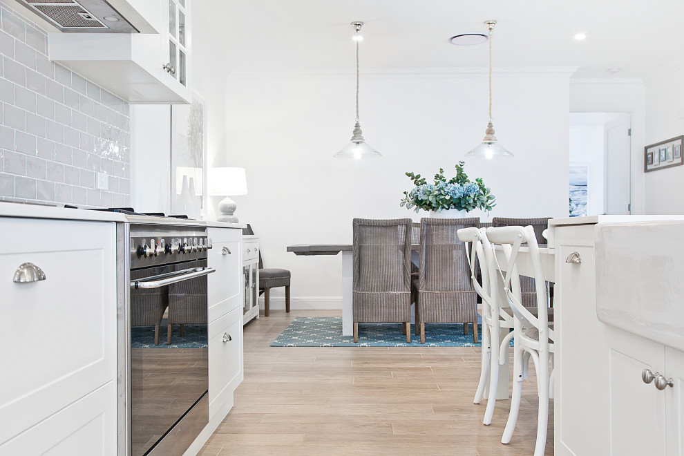 This is an example of a beach style kitchen in Other with a farmhouse sink, shaker cabinets, white cabinets, granite benchtops, grey splashback, ceramic splashback, stainless steel appliances, medium hardwood floors, with island, brown floor and white benchtop.