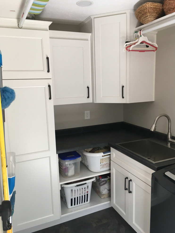 Photo of a mid-sized transitional l-shaped utility room in Other with a single-bowl sink, recessed-panel cabinets, white cabinets, laminate benchtops, grey walls, a side-by-side washer and dryer, black floor and black benchtop.