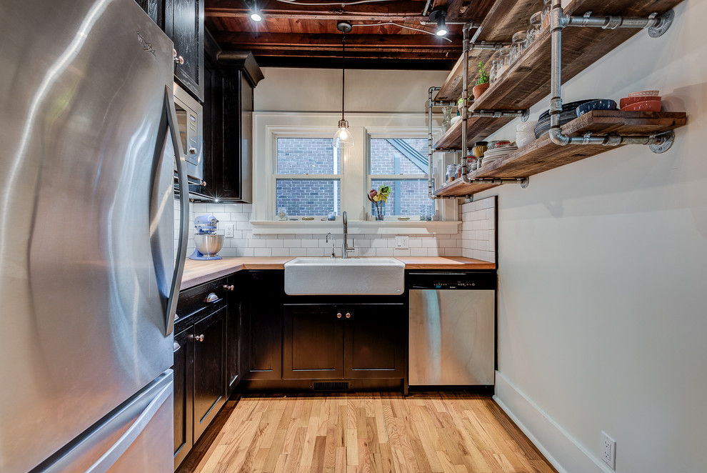 Small industrial u-shaped eat-in kitchen in Chicago with a farmhouse sink, shaker cabinets, black cabinets, wood benchtops, white splashback, stainless steel appliances, light hardwood floors, a peninsula and subway tile splashback.