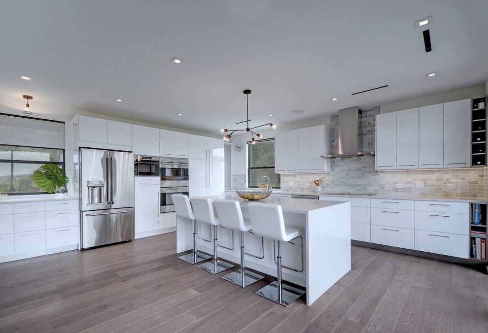 Contemporary l-shaped kitchen in Austin with flat-panel cabinets, white cabinets, grey splashback, subway tile splashback, stainless steel appliances, medium hardwood floors, with island, grey floor and white benchtop.