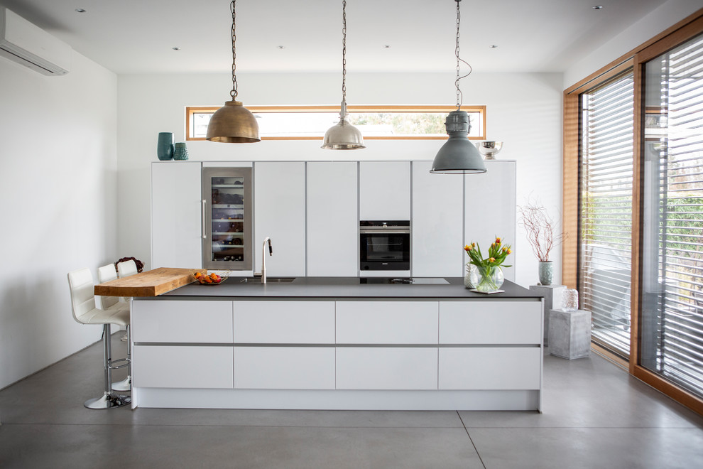 This is an example of a contemporary galley kitchen in Frankfurt with an undermount sink, flat-panel cabinets, white cabinets, black appliances, concrete floors, with island, grey floor and black benchtop.