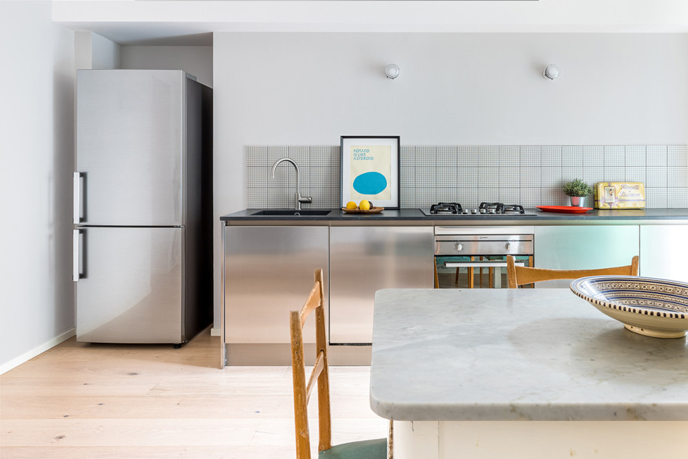 Mid-sized midcentury single-wall eat-in kitchen in Turin with an integrated sink, flat-panel cabinets, stainless steel cabinets, quartzite benchtops, grey splashback, ceramic splashback, stainless steel appliances, light hardwood floors, no island and black benchtop.
