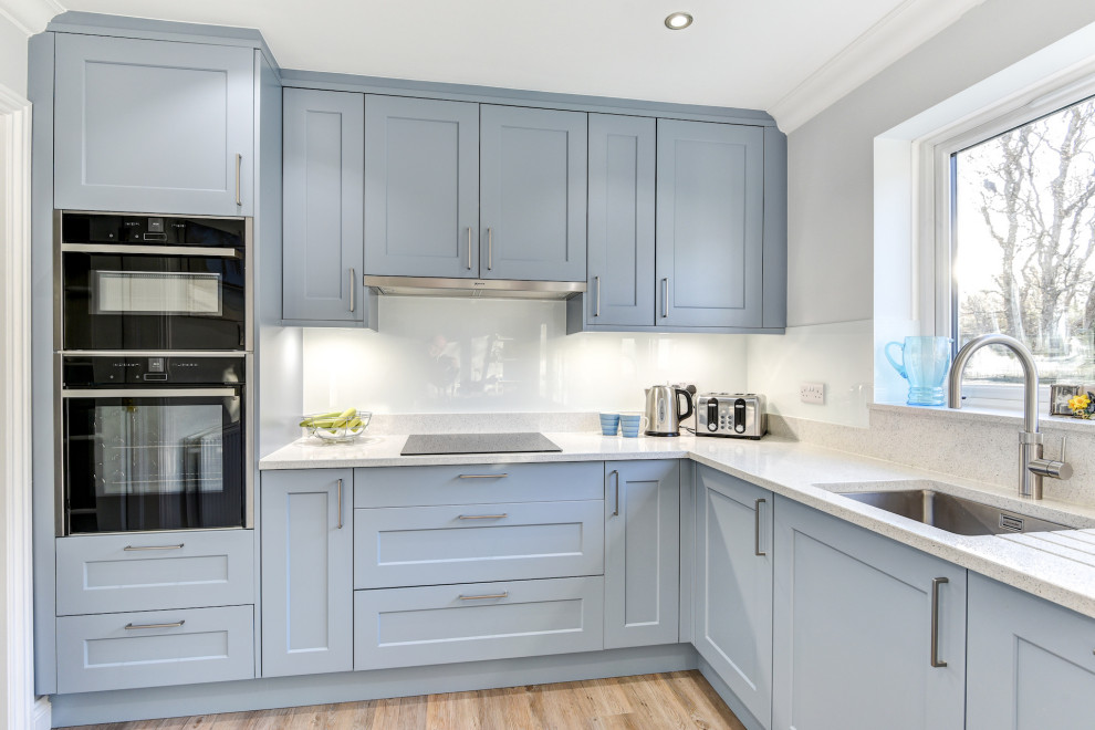 Exempel på ett mellanstort modernt vit vitt kök, med en undermonterad diskho, skåp i shakerstil, blå skåp, bänkskiva i kvartsit, vitt stänkskydd, glaspanel som stänkskydd, svarta vitvaror, ljust trägolv och brunt golv