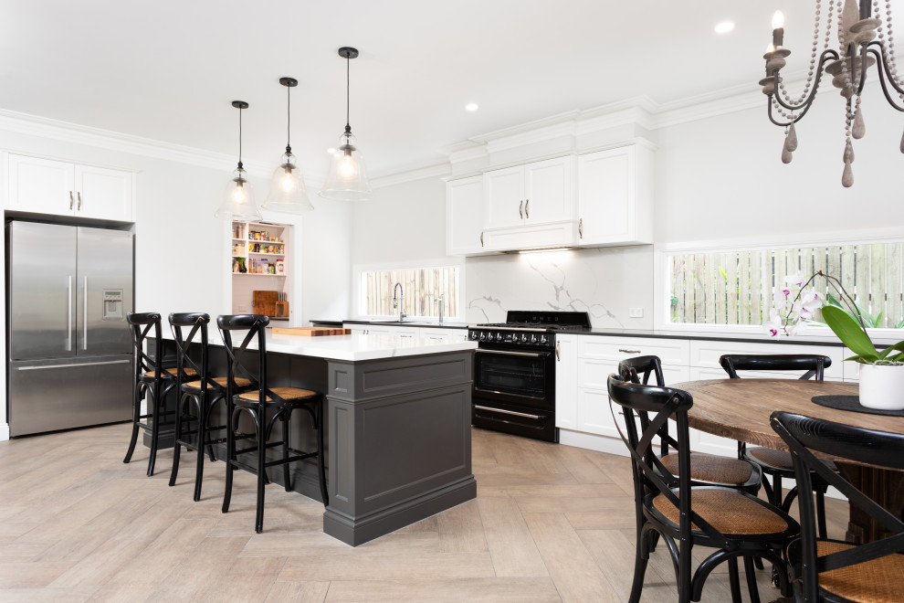 This is an example of a large beach style open plan kitchen in Brisbane with an undermount sink, shaker cabinets, white cabinets, granite benchtops, stone slab splashback, black appliances, ceramic floors, with island, brown floor and black benchtop.