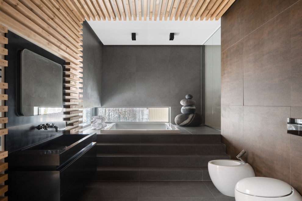 Inspiration for a country master bathroom with flat-panel cabinets, black cabinets, a japanese tub, a wall-mount toilet, brown tile, gray tile, a vessel sink and grey floor.