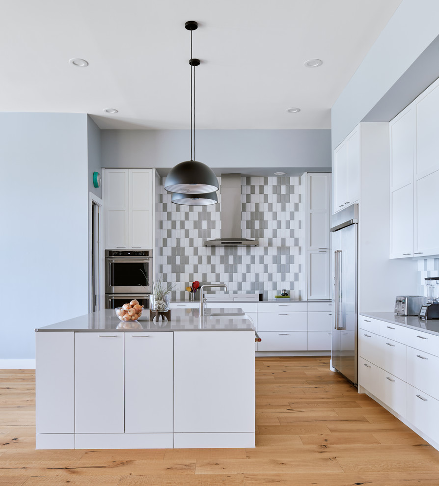 Photo of a contemporary kitchen in Austin with a farmhouse sink, flat-panel cabinets, white cabinets, multi-coloured splashback, stainless steel appliances, light hardwood floors, with island, beige floor and grey benchtop.