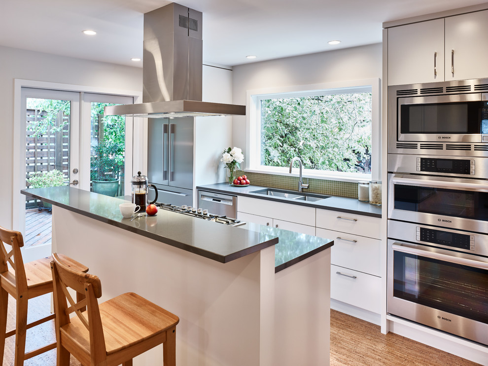 This is an example of a small contemporary galley kitchen in Vancouver with an undermount sink, flat-panel cabinets, white cabinets, quartz benchtops, green splashback, glass tile splashback, stainless steel appliances, cork floors and with island.