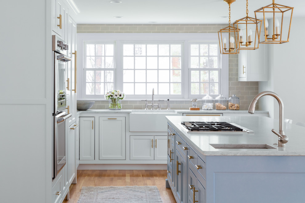 Transitional l-shaped kitchen in Boston with a farmhouse sink, recessed-panel cabinets, white cabinets, beige splashback, subway tile splashback, stainless steel appliances, medium hardwood floors, with island, brown floor and white benchtop.