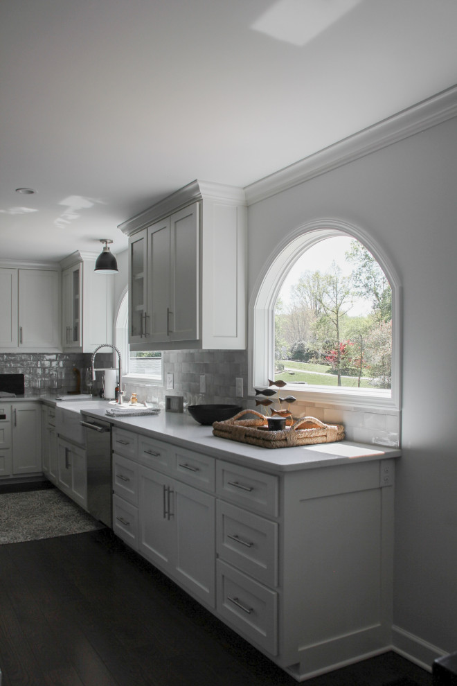 This is an example of a small beach style l-shaped eat-in kitchen in Charlotte with a farmhouse sink, shaker cabinets, white cabinets, quartz benchtops, grey splashback, ceramic splashback, stainless steel appliances, dark hardwood floors, with island, brown floor and white benchtop.