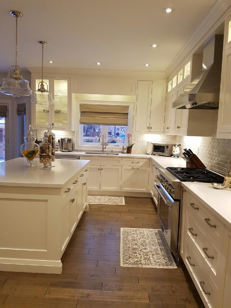 Open concept kitchen - mid-sized transitional l-shaped medium tone wood floor and brown floor open concept kitchen idea in Toronto with an undermount sink, beaded inset cabinets, white cabinets, solid surface countertops, white backsplash, brick backsplash, stainless steel appliances, an island and white countertops
