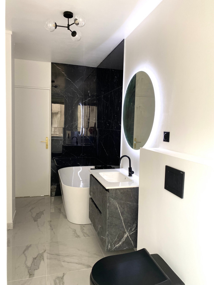 Foto på ett stort funkis vit en-suite badrum, med svarta skåp, ett fristående badkar, en vägghängd toalettstol, svart kakel, marmorkakel, vita väggar, marmorgolv, bänkskiva i akrylsten och vitt golv