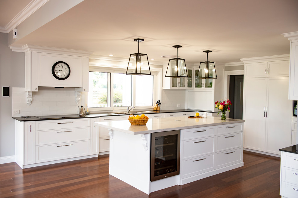 Large traditional u-shaped kitchen pantry with a double-bowl sink, shaker cabinets, white cabinets, marble benchtops, white splashback, ceramic splashback, black appliances, dark hardwood floors, with island, brown floor and black benchtop.