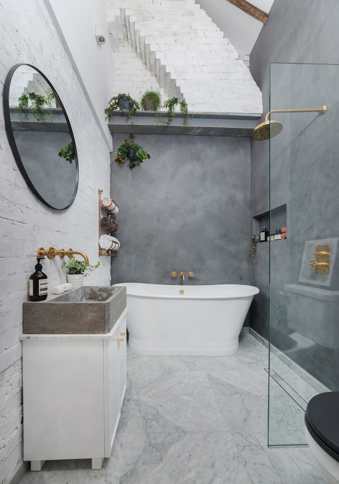 This is an example of a mid-sized industrial bathroom in London with a freestanding tub, an open shower, white walls, marble floors, white cabinets, a one-piece toilet, a trough sink, marble benchtops, multi-coloured floor and an open shower.